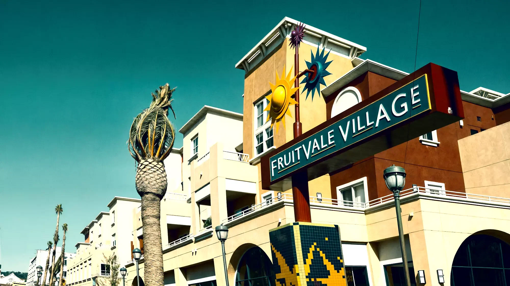 Daytime scene of Fruitvale Village with a multi-story building, palm tree, and colorful sign featuring sun motifs, reflecting the vibrant atmosphere of East Bay.