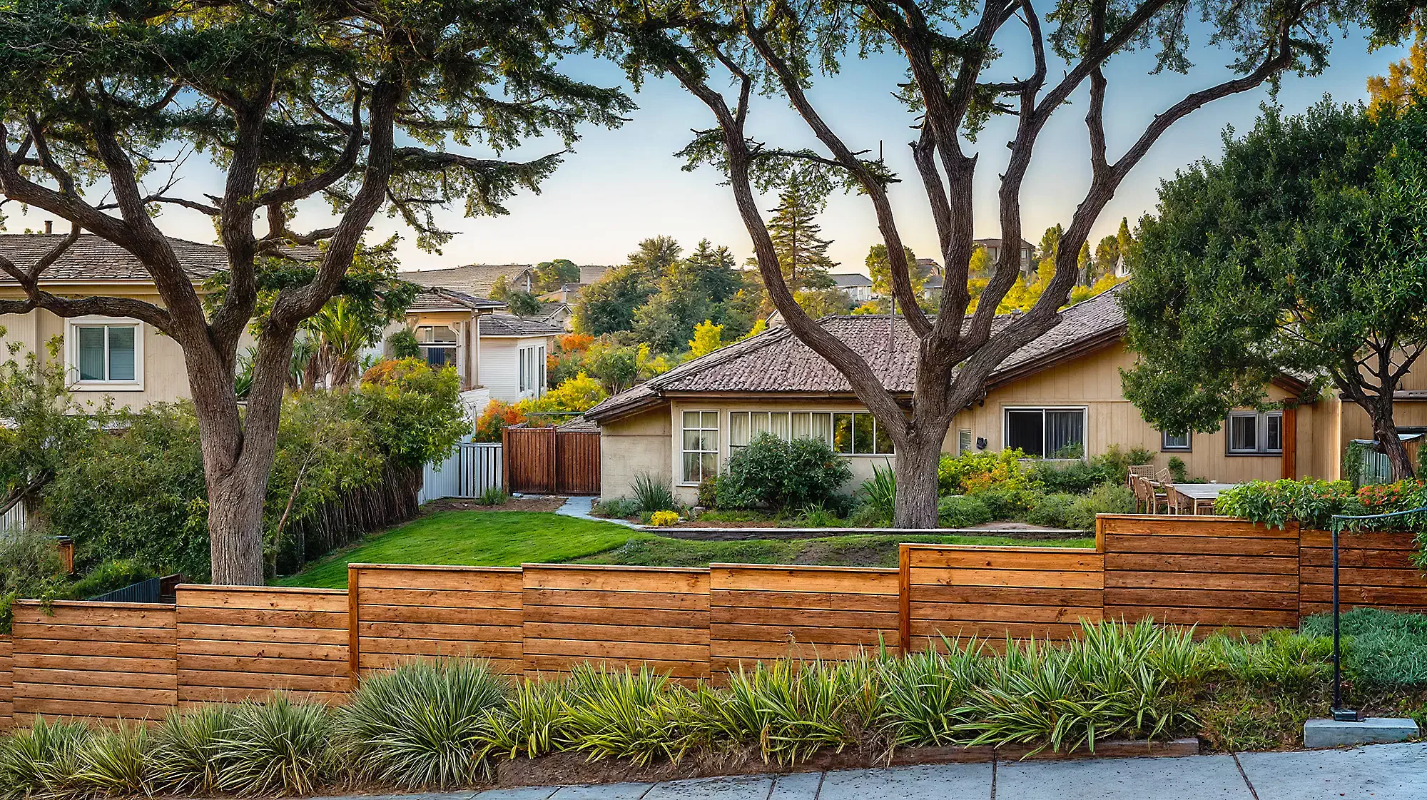 A suburban scene with a wooden fence in the foreground, a green lawn, and houses managed by rental property management companies surrounded by large trees and shrubs in the background.