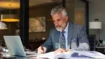 A man in a suit sits at a desk, writing on papers with a pen, while looking at a laptop in an office setting.