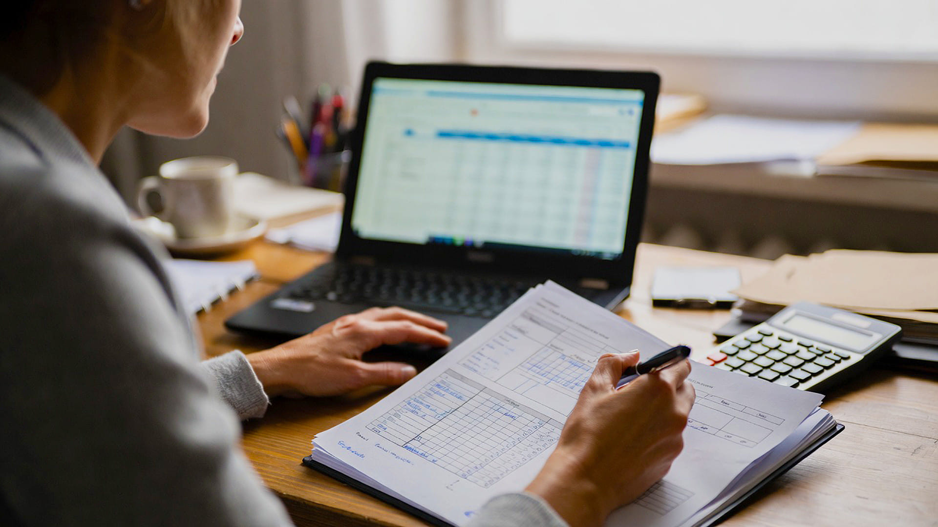Person reviewing printed financial documents and writing notes at a desk with an open laptop displaying a spreadsheet, calculator, and coffee cup nearbyโdemonstrating the precision required for professional property management bookkeeping services.