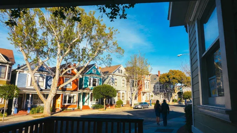 A row of colorful houses on a sunny residential street, managed by SLPM Alameda California Property Management, with two people walking on the sidewalk under a clear blue sky.