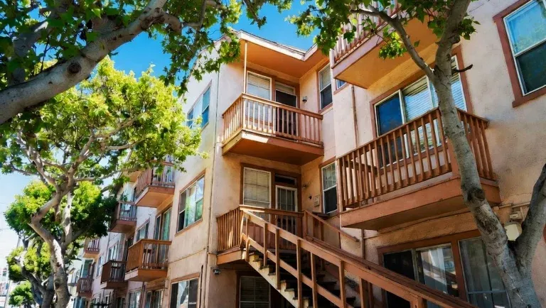 Three-story apartment building with peach-colored walls, wooden balconies and stairs, and leafy trees in front under a clear blue skyโprofessionally cared for by SLPM Oakland Property Management.