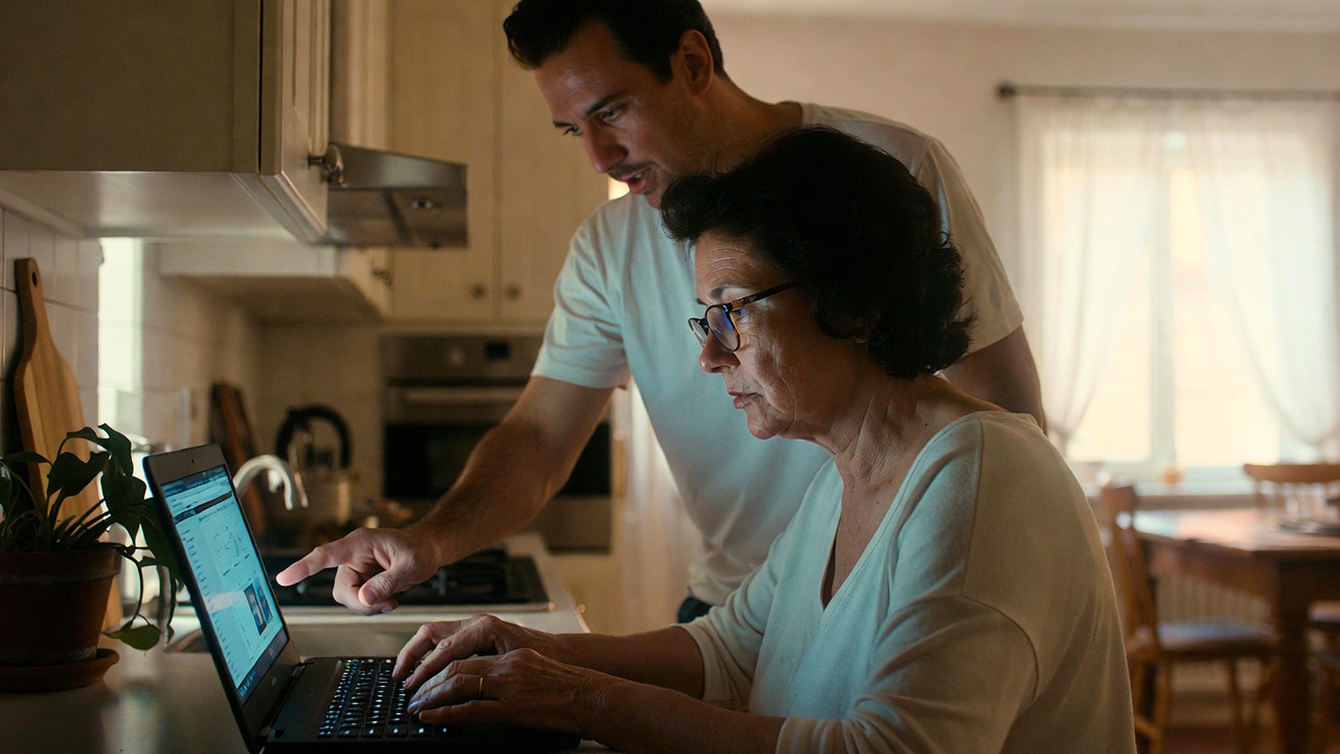 A woman uses a laptop in a kitchen while a man stands beside her, pointing at the screen and providing assistance—perhaps reviewing Bay Area Property Management listings together.