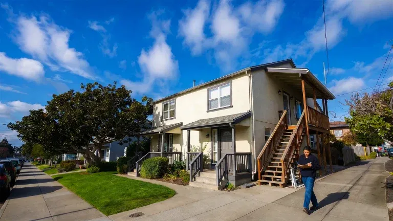 Two-story beige apartment building with exterior stairs, green lawn, trees, and blue sky. A person walks on the sidewalk in front—ideal for landlords or a property manager seeking a well-maintained residence.