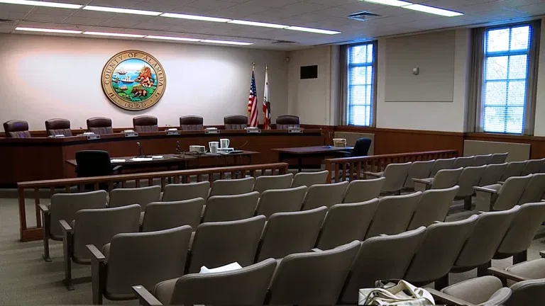 Empty government meeting room with rows of chairs, a long desk with microphones, American and state flags, and a county seal on the wall—ideal for gatherings or presentations managed by SLPM Bay Area Property Management.