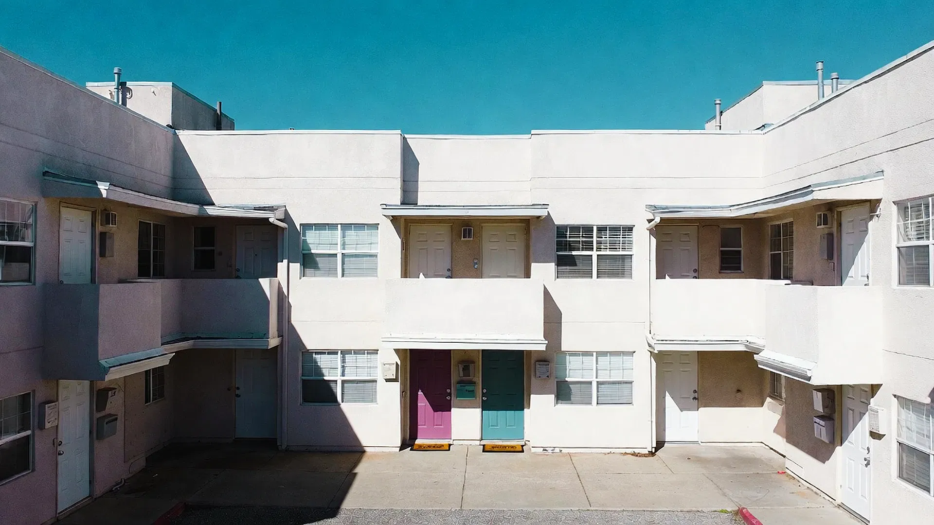 A two-story apartment building with white walls, flat roof, and two central doors—one pink, one blue—managed by SLPM Bay Area Property Management, stands under a clear, bright blue sky.