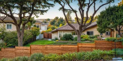 A suburban scene with a wooden fence in the foreground, a green lawn, and houses managed by rental property management companies surrounded by large trees and shrubs in the background.